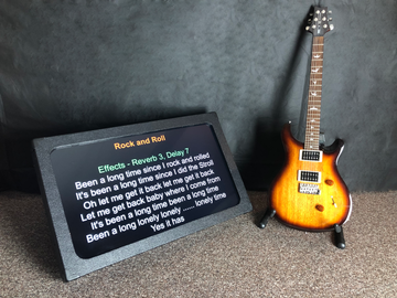 Teleprompter screen displaying song lyrics next to an electric guitar on a carpeted floor.